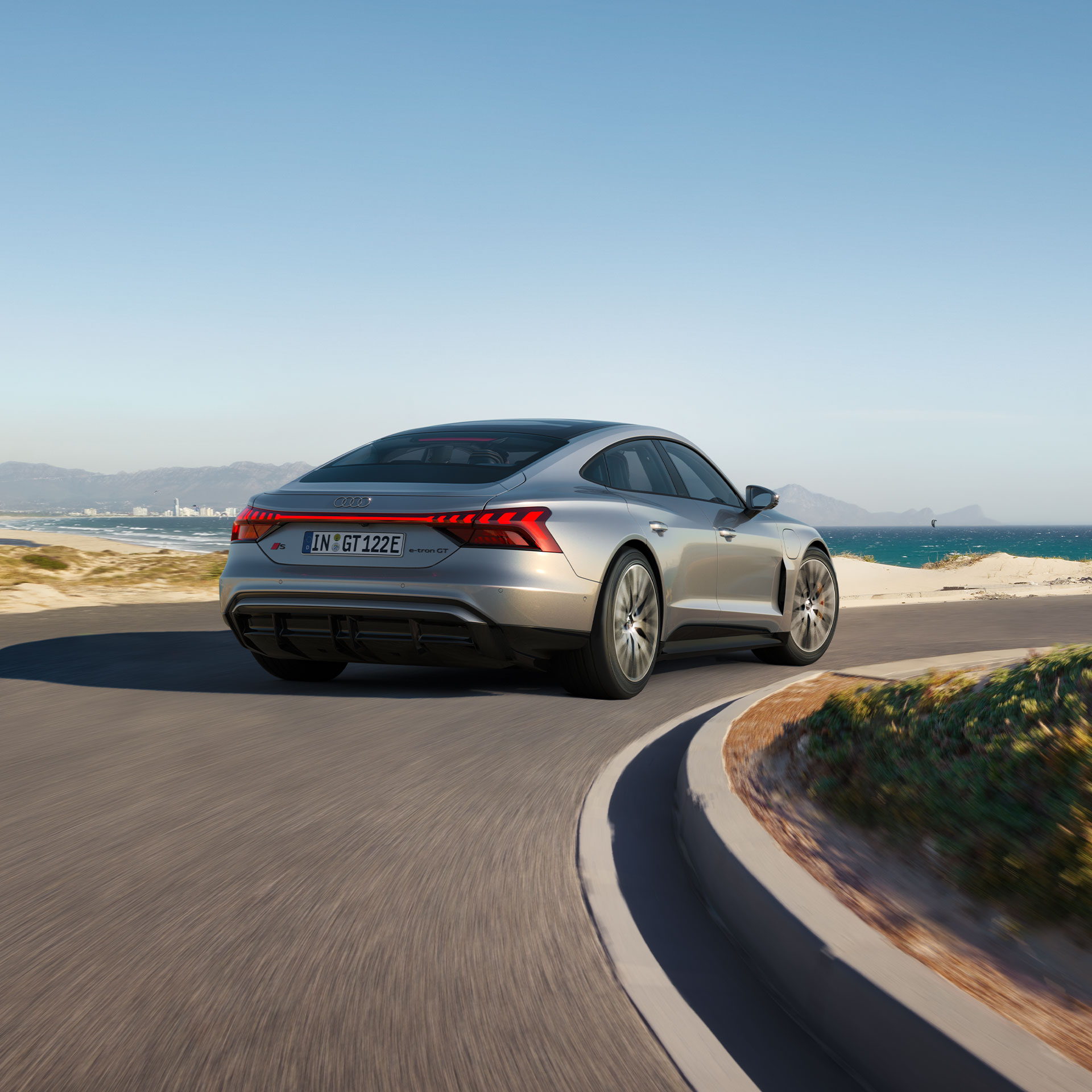  A silver Audi S e-tron GT drives along a winding coastal road with a view of the sea and a city in the background.